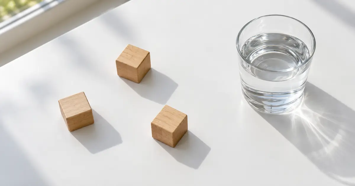 昼下がりの窓辺の白いテーブルに、三つの小さな木のさいころとガラスのコップの水を真上から見たところ