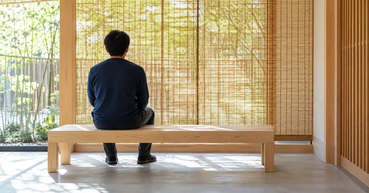 昼の光が差し込む和の待合で、すだれの前の白木のベンチに座ってくつろぐ、紺のセーターの男性の後ろ姿
