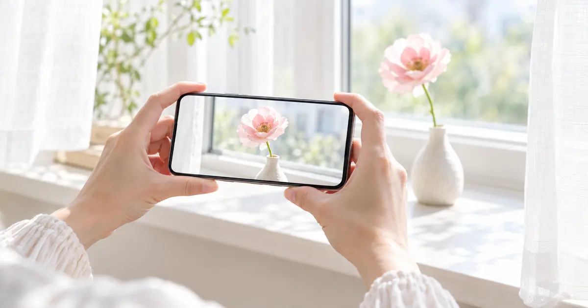 窓辺に置いた一輪挿しの花を、スマートフォンのカメラで撮っている手元