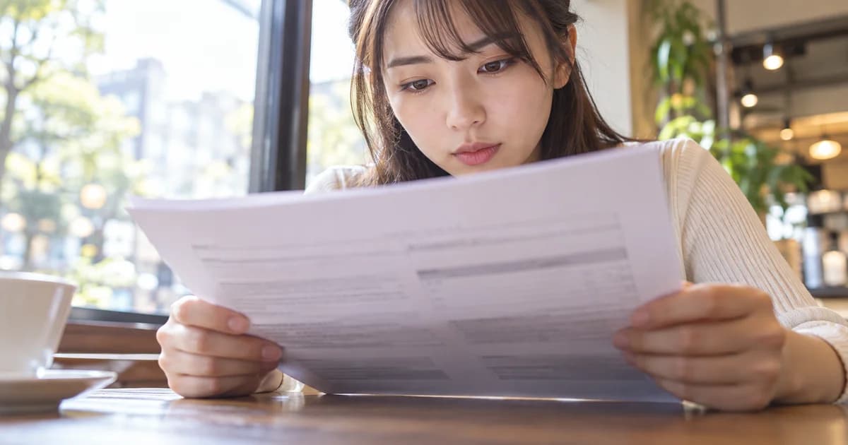 明るいカフェの窓際の席で、契約書の紙を両手で持ち、文面を読み込んでいる女性を、テーブルの向かい側の低い位置から