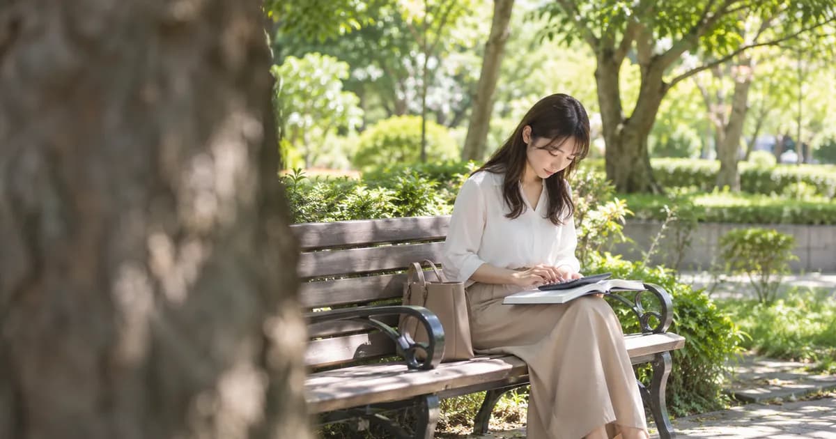 晴れた休日の公園のベンチで、膝に広げたノートの上で電卓をたたいている女性を、手前の木の幹越しに少し離れて